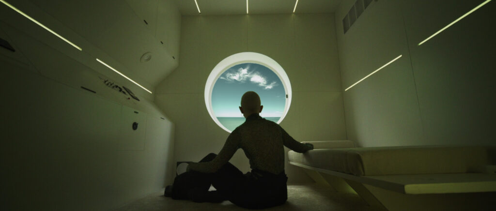 Still from MESSIS by Van Grimde Corps Secrets — a woman gazes through a porthole toward the sky from a stark white cell